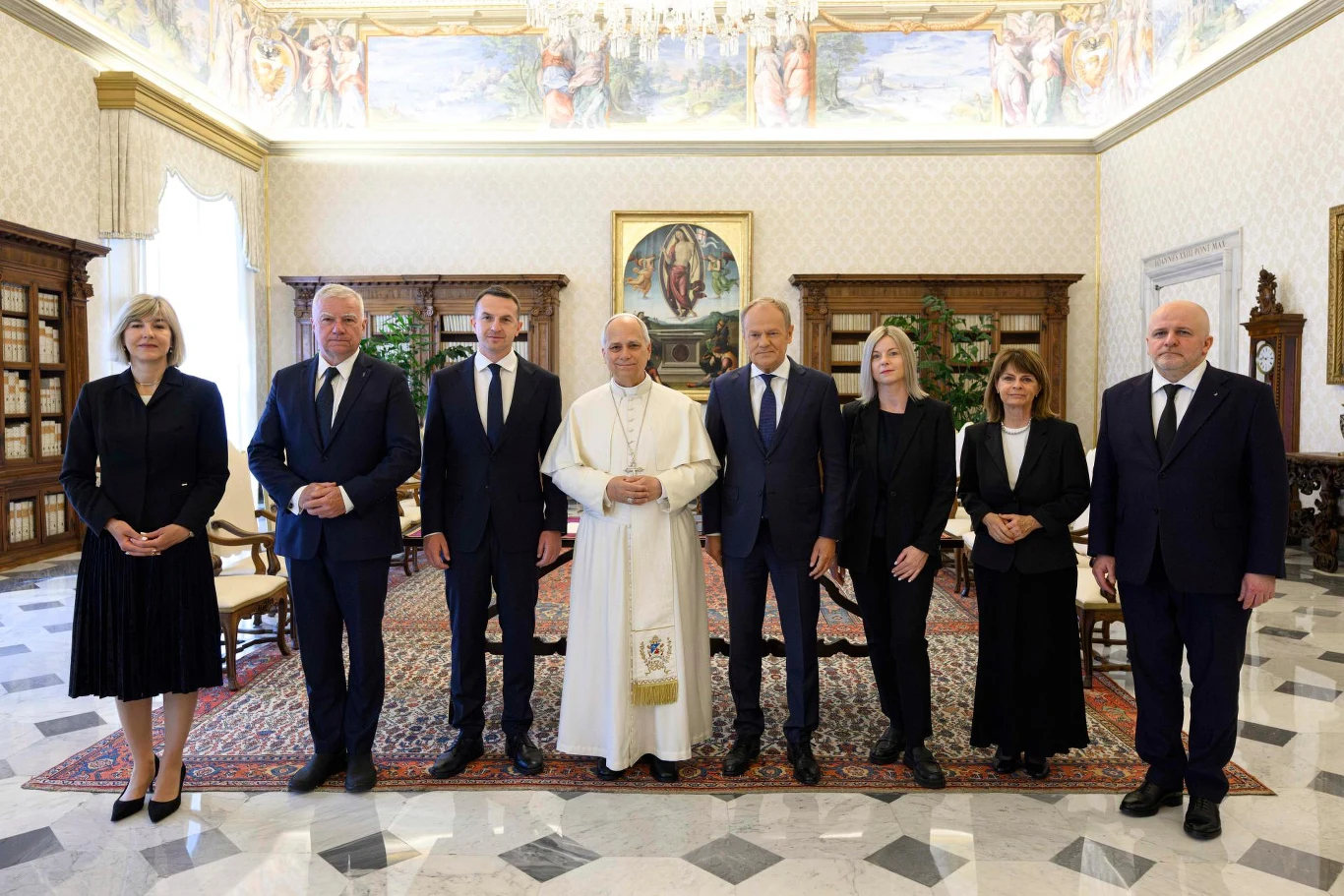 Grupa ośmiu elegancko ubranych osób stoi w rzędzie w ozdobnym, pałacowym wnętrzu, w centrum postać w białej szacie z krzyżem na piersi.