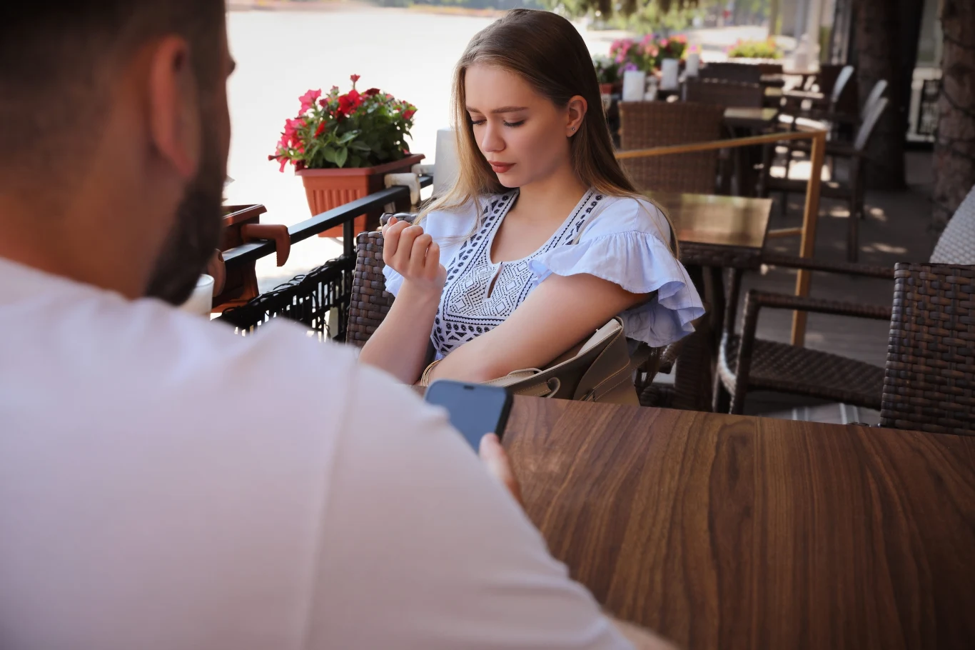 Młoda kobieta siedzi przy stoliku w kawiarni na tarasie, patrząc na swoje paznokcie, podczas gdy naprzeciwko niej mężczyzna używa telefonu komórkowego.