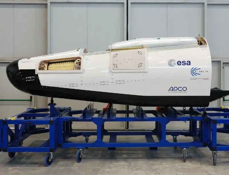 Moduł testowy europejskiego miniwahadłowca kosmicznego z charakterystycznymi oznaczeniami ESA i APCO, umieszczony na niebieskim stojaku technicznym w hali montażowej.