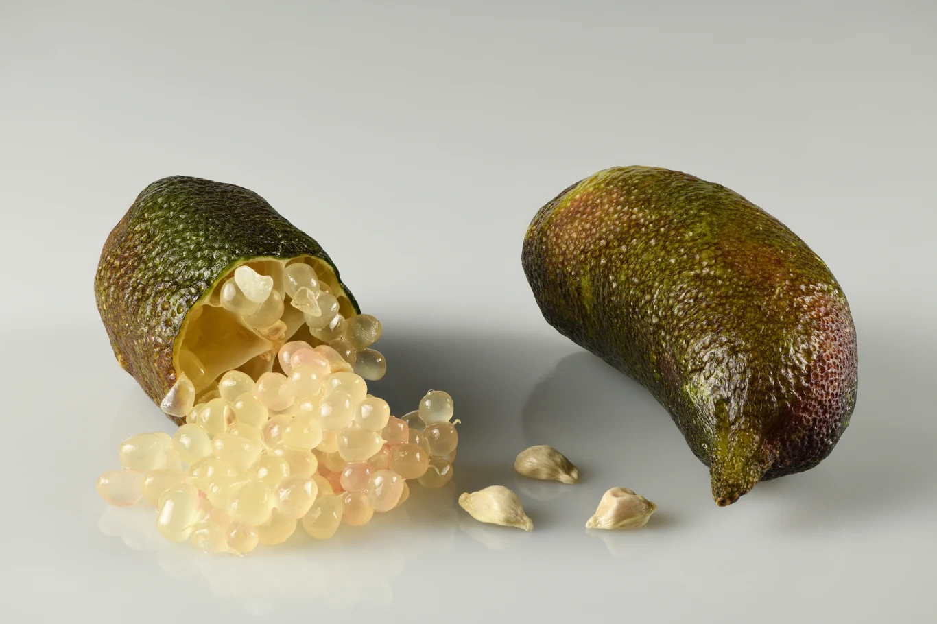 Po przekrojeniu z finger lime wysypują się drobne kuleczki przypominające kawior Dwa owoce limonkowego kawioru, jeden przekrojony z widocznymi przezroczystymi, kulistymi ziarenkami i kilkoma jasnymi pestkami obok.