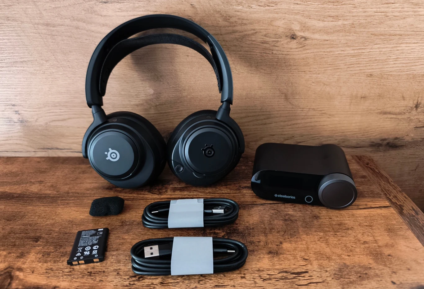 Czarne słuchawki bezprzewodowe z mikrofonem i logiem SteelSeries, stacja bazowa, dwa przewody USB, mikrofon z gąbką oraz wymienna bateria ułożone na drewnianej powierzchni.