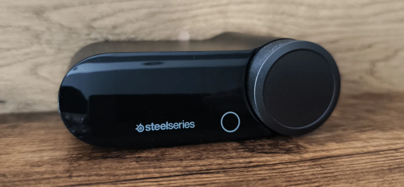 Czarny kontroler Steelseries z dużym pokrętłem i podświetlanym przyciskiem na drewnianej powierzchni.