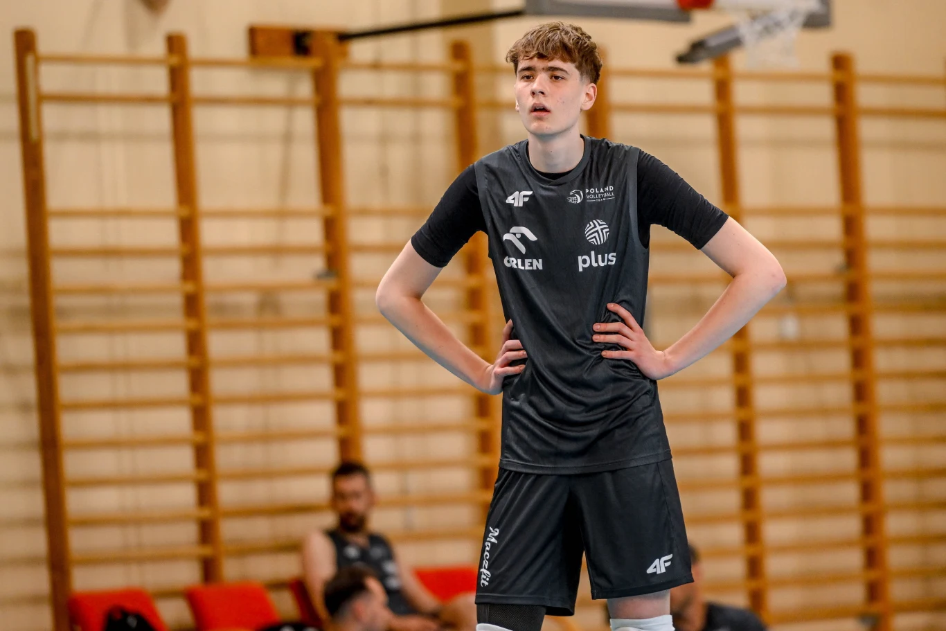Młody siatkarz w czarnym stroju sportowym z logotypami sponsorów stoi z rękami na biodrach na tle sali gimnastycznej z drewnianą drabinką.