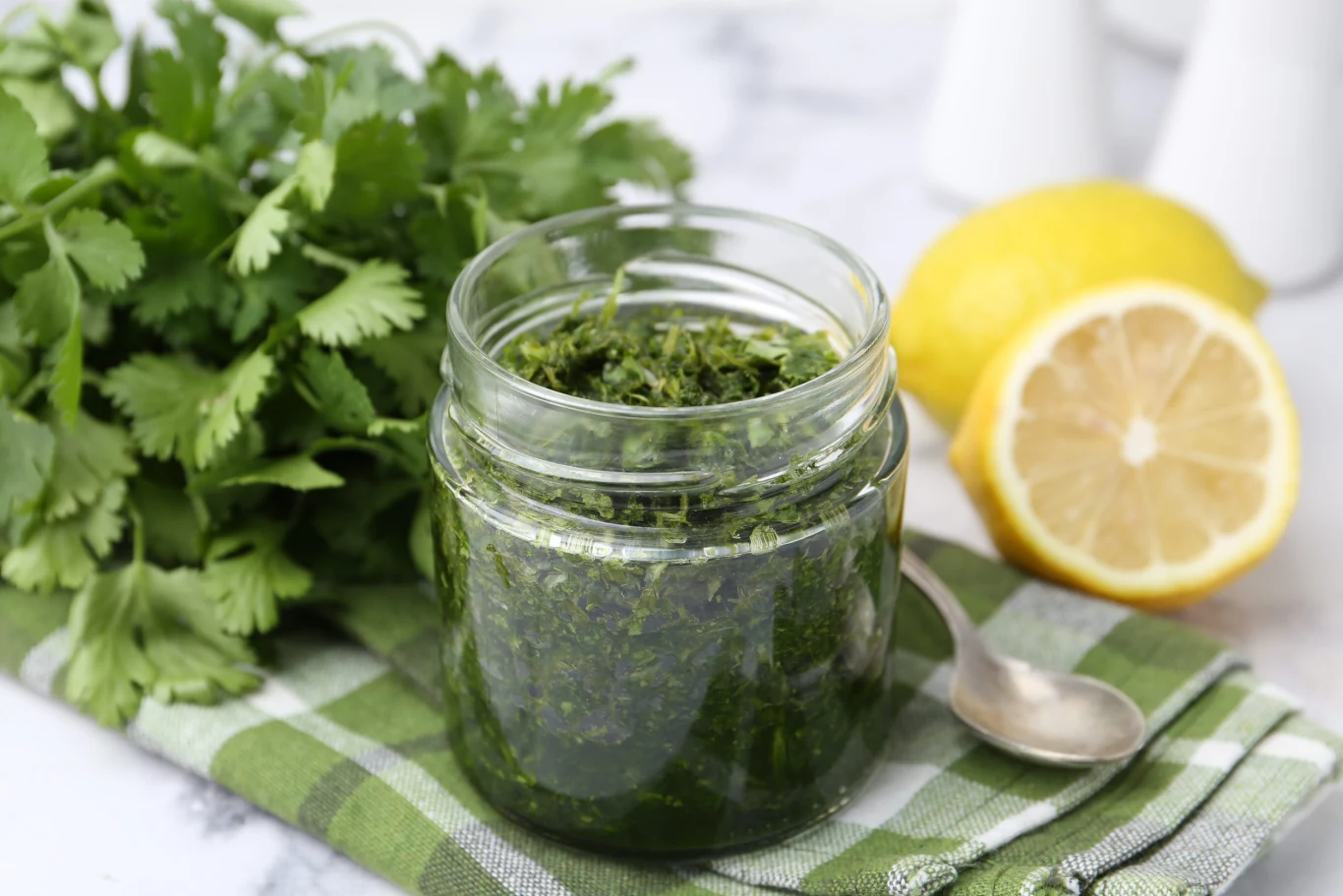 Słoik z zielonym pesto ustawiony na zielono-białej ściereczce, obok świeżej natki kolendry i przekrojonej cytryny.