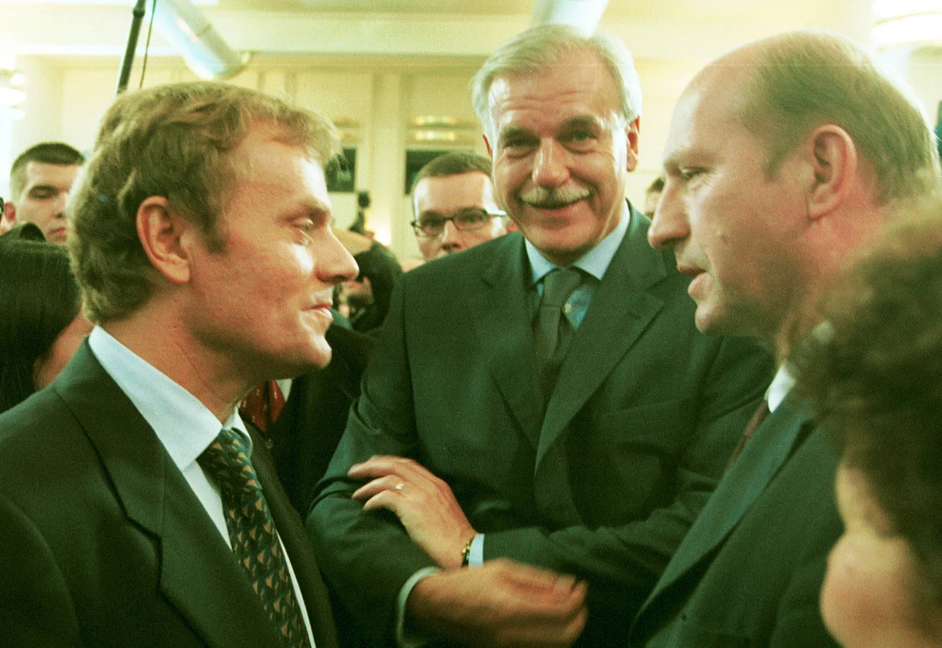 Trzech elegancko ubranych mężczyzn w garniturach prowadzi rozmowę w jasnym, oficjalnym pomieszczeniu pełnym ludzi.
