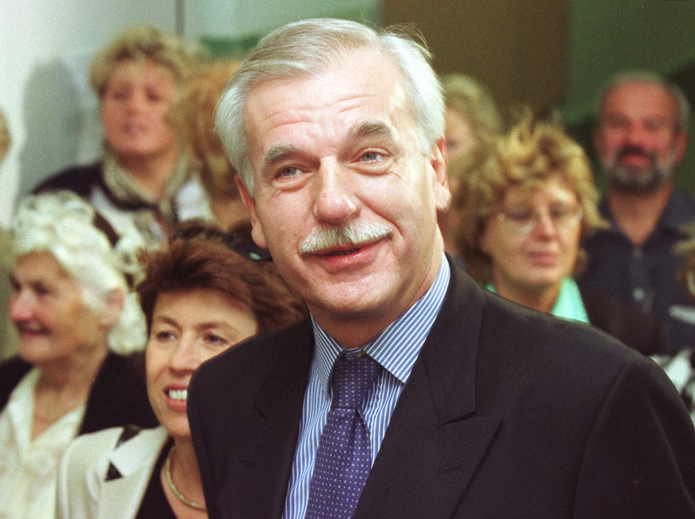 Uśmiechnięty mężczyzna z siwymi włosami i wąsem w garniturze, otoczony grupą osób w różnym wieku, większość z nich patrzy w jego stronę.