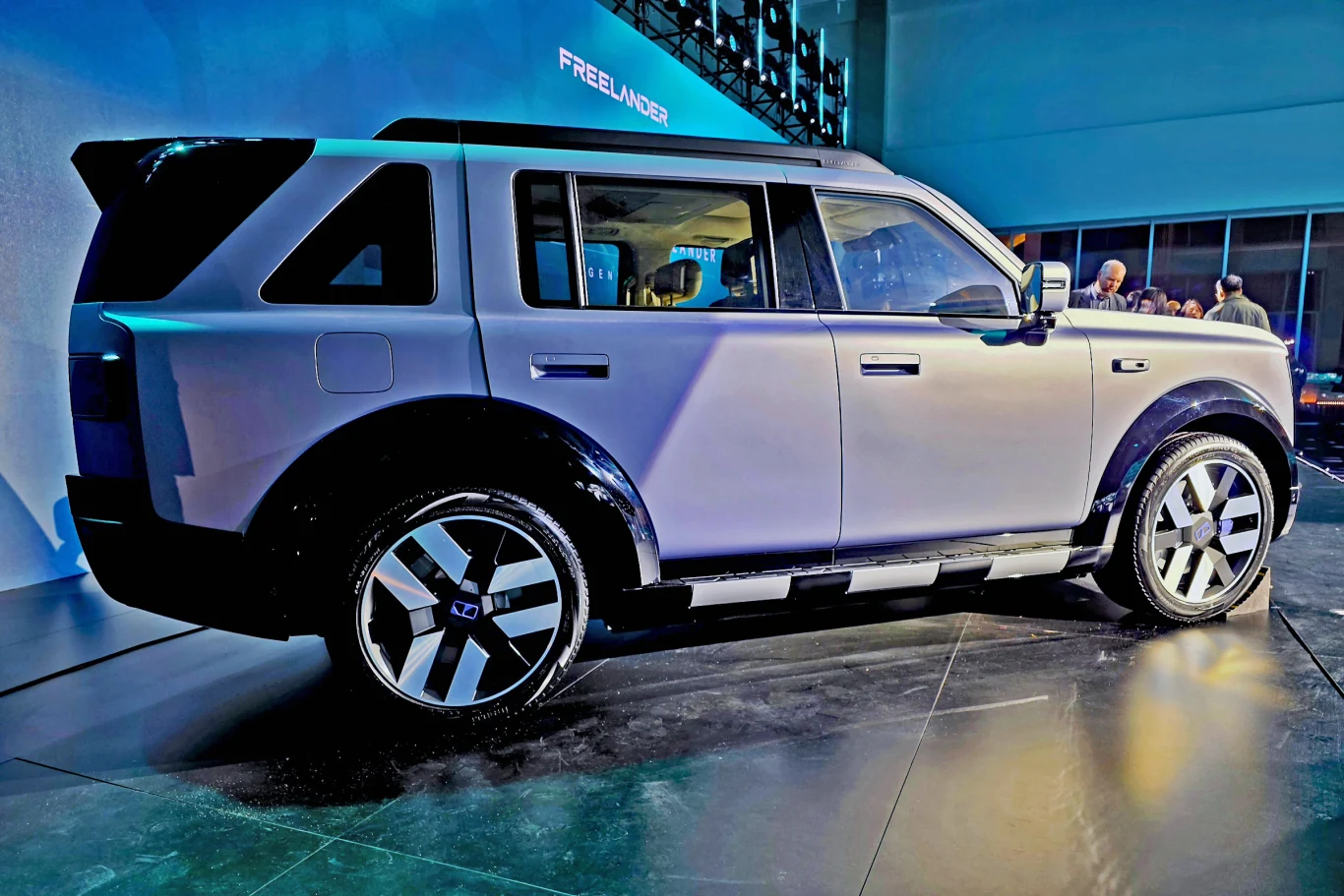 Nowoczesny SUV o kanciastych liniach nadwozia i dużych felgach, ustawiony na ekspozycji wewnętrznej w świetle reflektorów, z otwartymi drzwiami kierowcy i obecnością kilku osób obserwujących w tle.