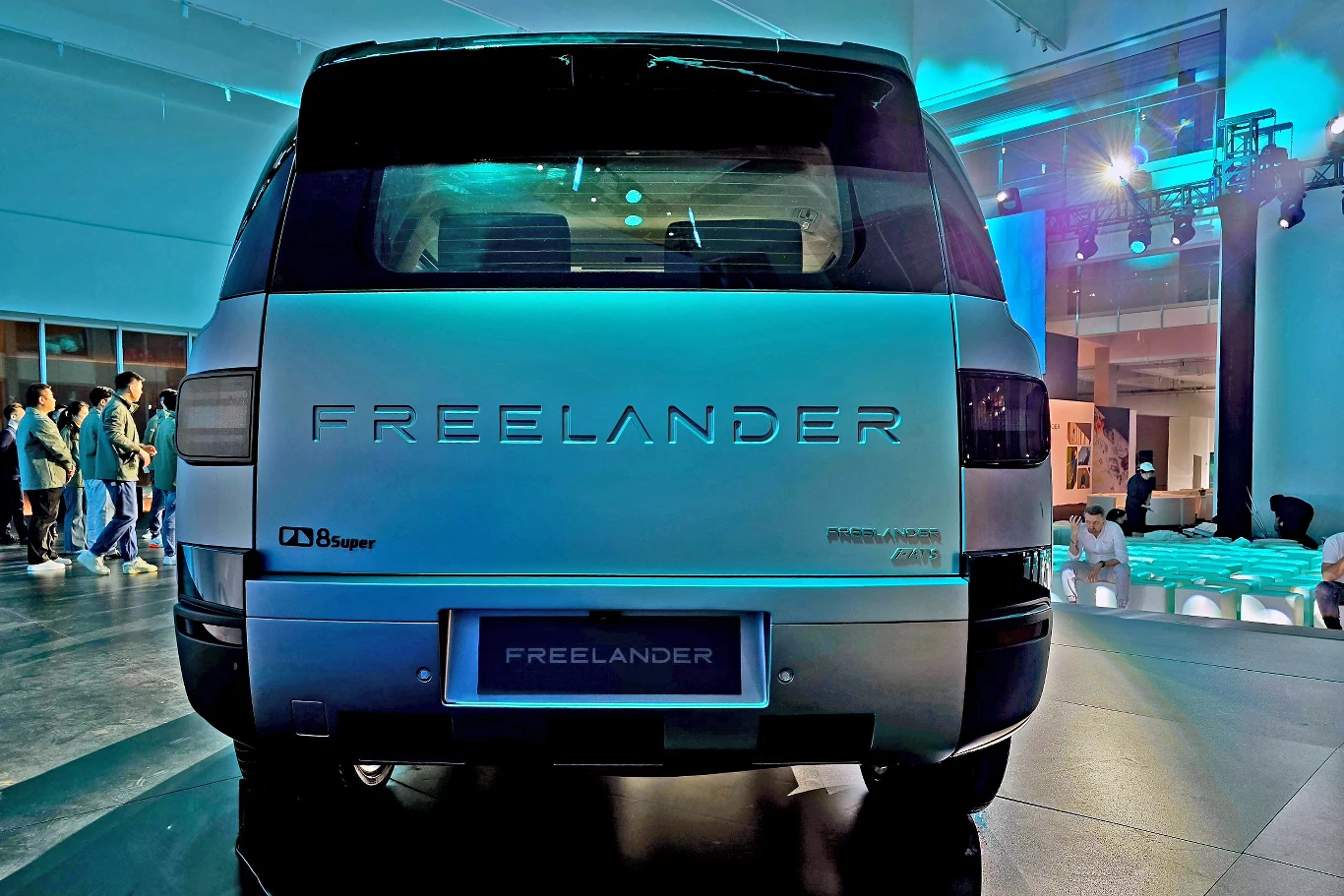 Tylna część jasnoszarego samochodu Freelander z wyraźnie widocznym logo oraz detalami świateł, stojący w nowoczesnym, oświetlonym wnętrzu wystawowym.