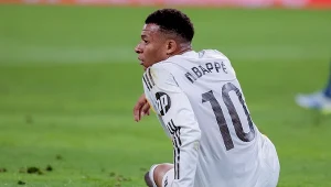 Zawodnik w białym stroju piłkarskim z numerem 10 i nazwiskiem „Mbappé” siedzi na murawie stadionu, spoglądając w bok.