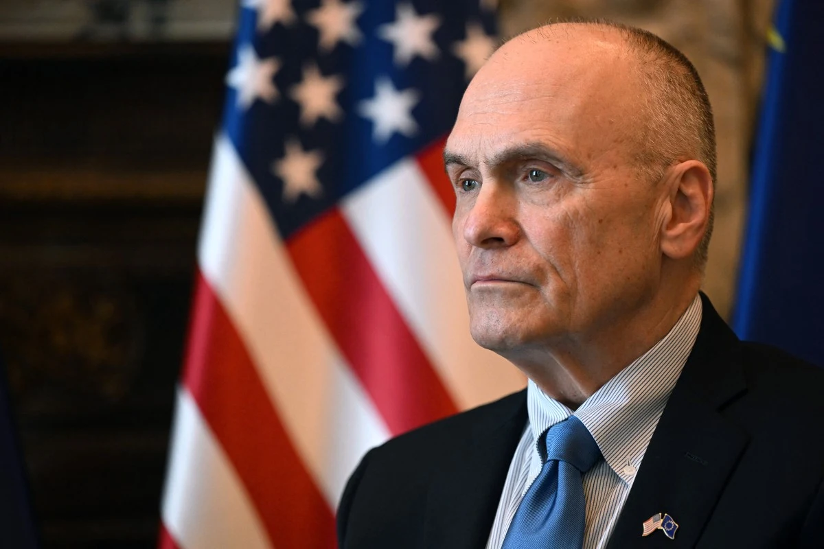 Mężczyzna w garniturze z niebieskim krawatem i przypiętą flagą USA, w tle flaga Stanów Zjednoczonych.
