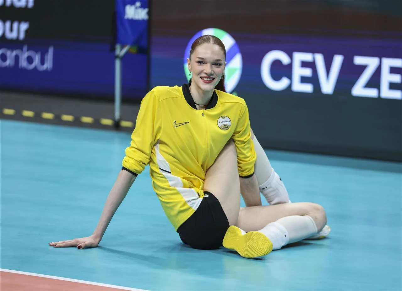 Uśmiechnięta siatkarka w żółtym stroju sportowym siedzi na niebieskim parkiecie hali, mając jedną nogę wyprostowaną, a drugą ugiętą, z logo klubu na piersi.