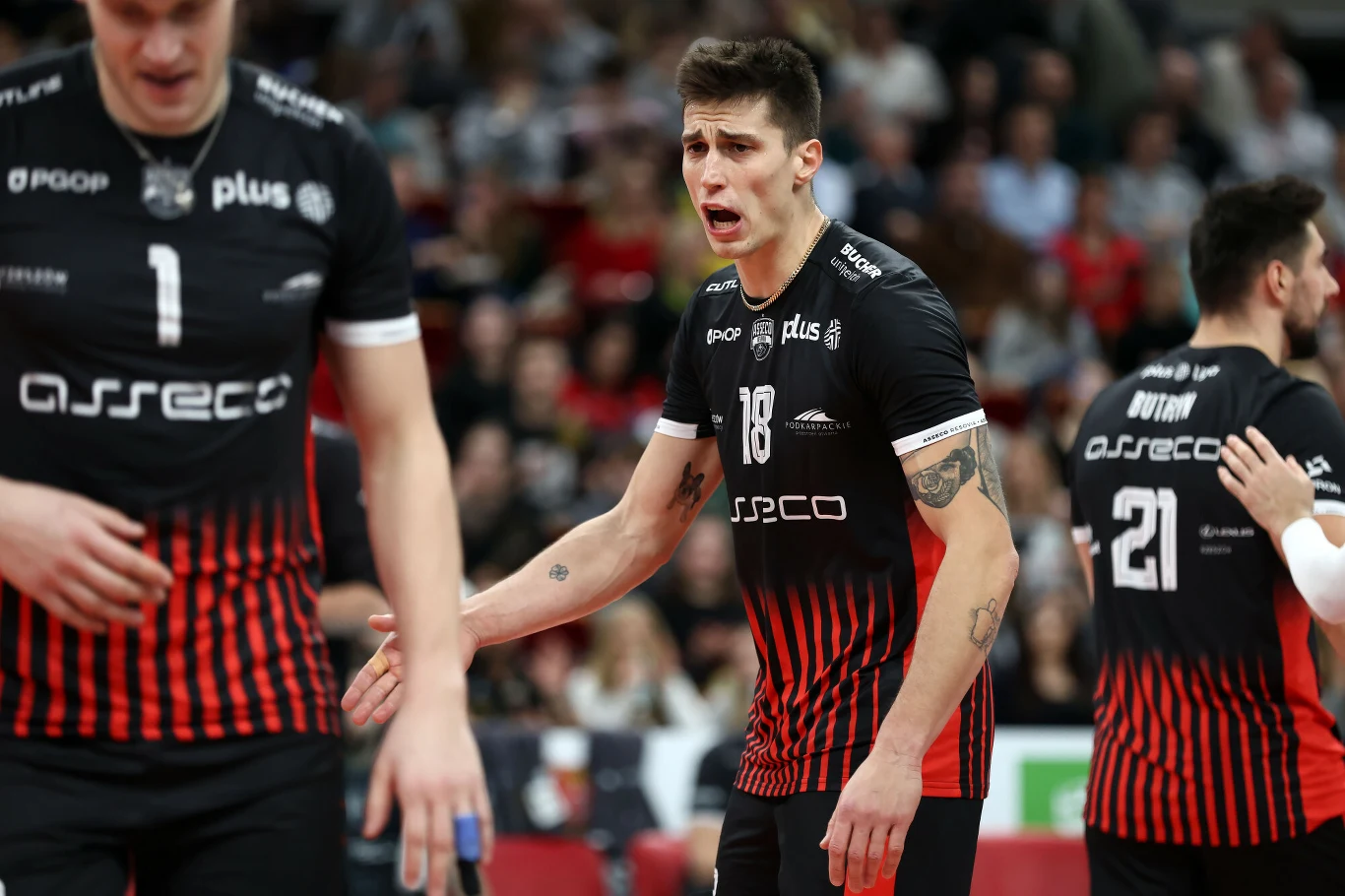 Siatkarz w czarno-czerwonym stroju z numerem 18 wyraża silne emocje podczas meczu, wokół niego znajdują się pozostali zawodnicy drużyny Asseco Resovia również w takich samych strojach.