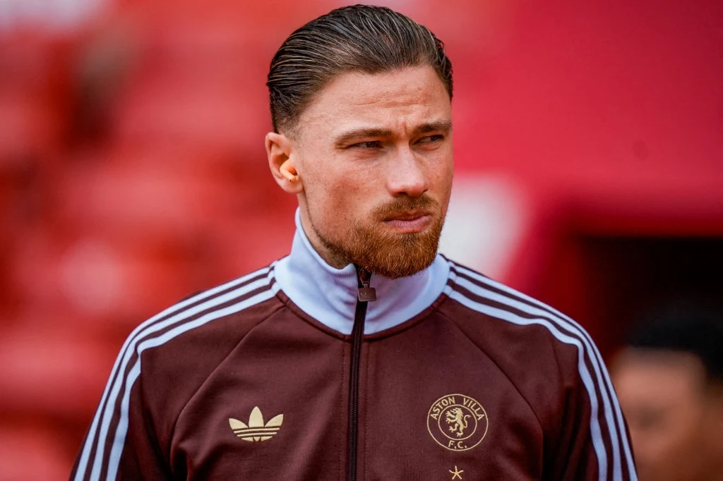Mężczyzna w bordowej sportowej kurtce z białymi paskami i logo drużyny Aston Villa, uczesany do tyłu z krótką brodą, stoi na tle czerwonego stadionu.