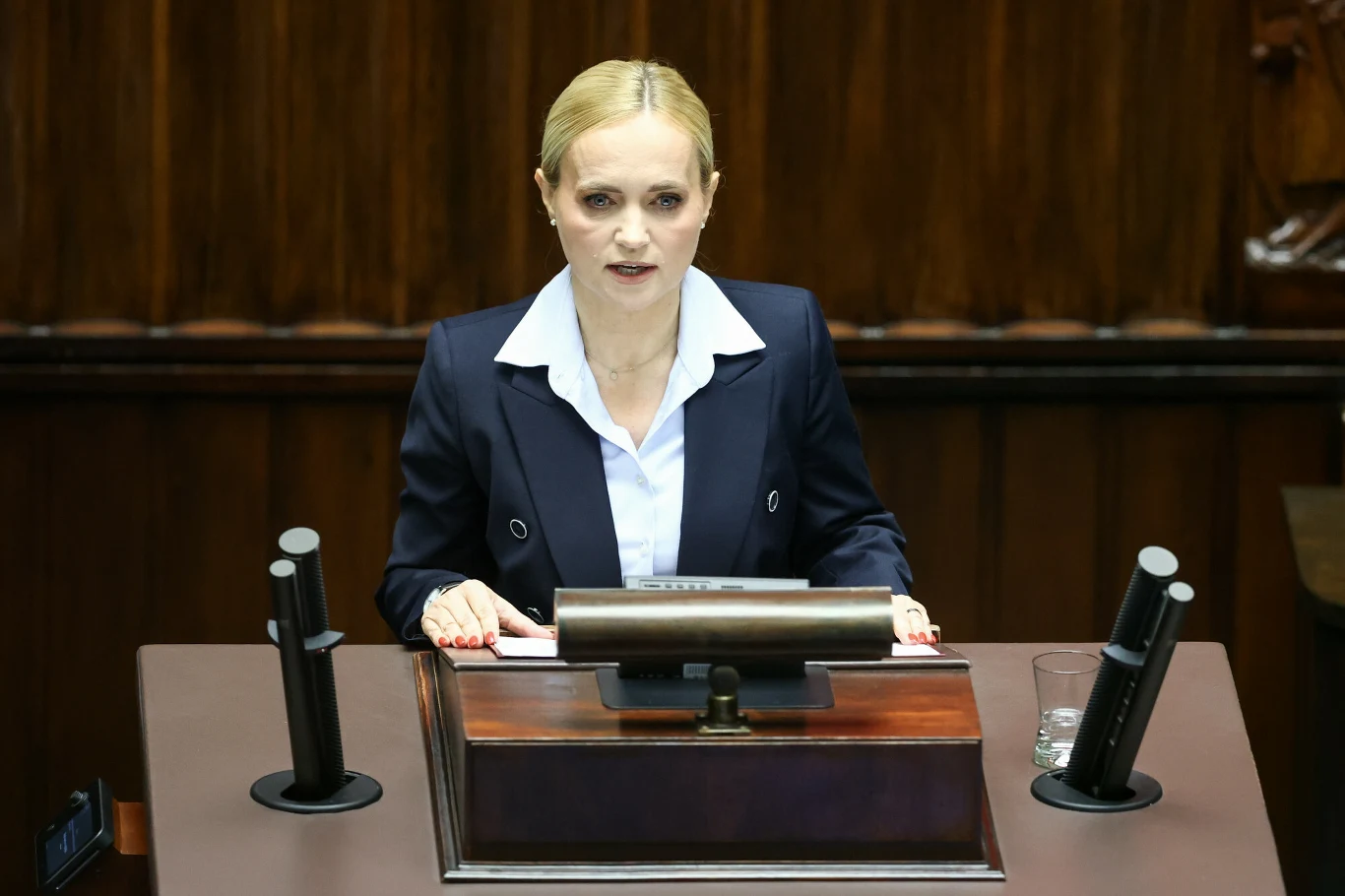 Minister zdrowia Jolanta Sobierańska-Grenda Jolanta Sobierańska-Grenda przemawia zza mównicy w sali obrad, ubrana w granatowy garnitur, mikrofony i szklanka wody.