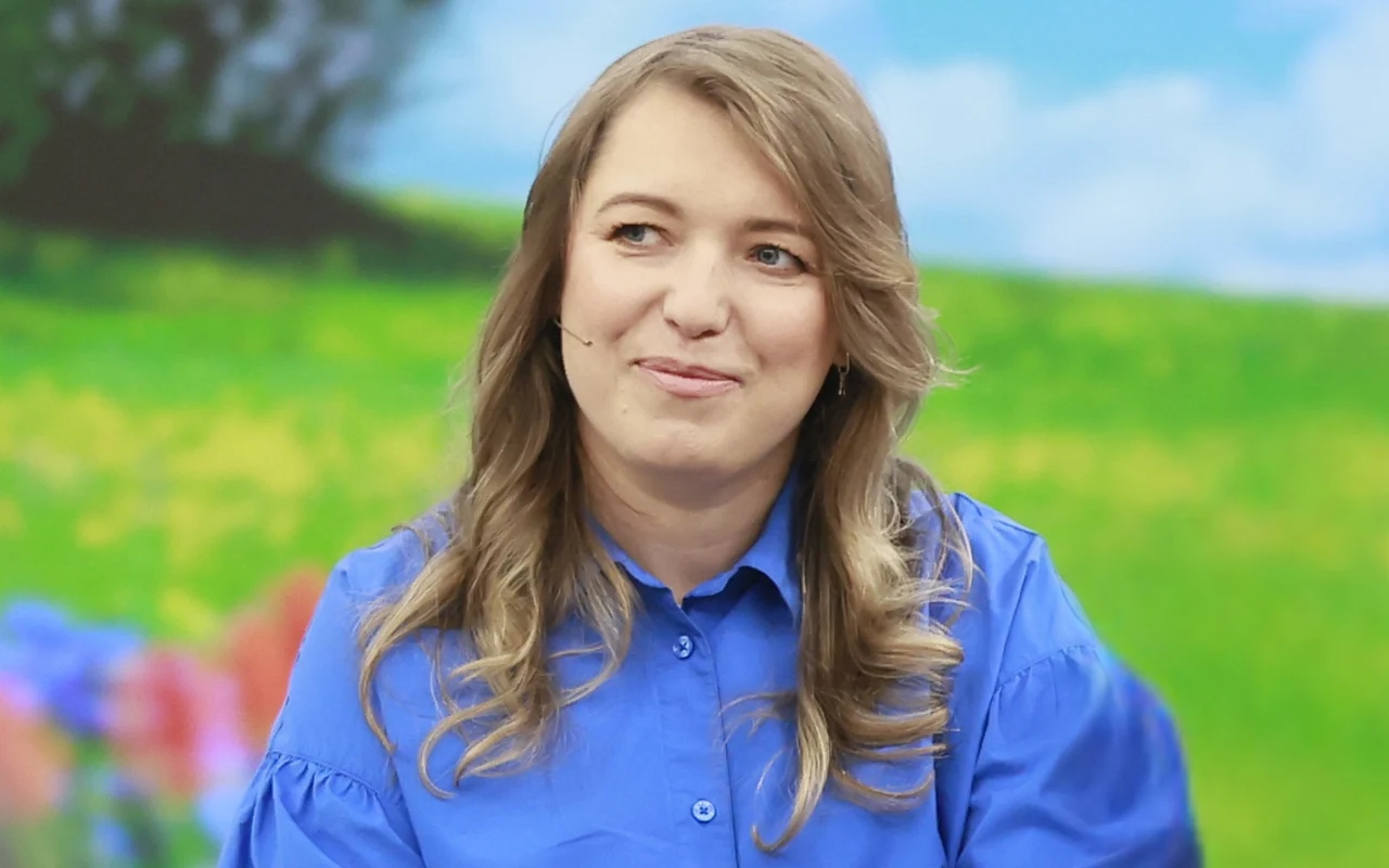 Kobieta w niebieskiej koszuli z mikrofonem na twarzy, siedząca na tle kolorowego, rozmytego krajobrazu z zieloną trawą i niebem.