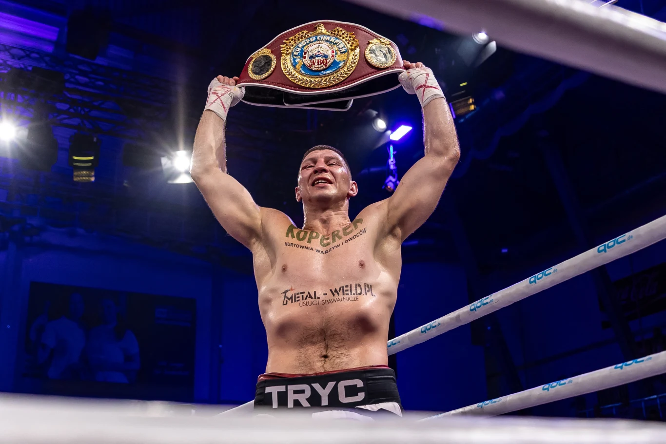 Bokser z uniesionymi rękami trzyma trofeum mistrzowskie nad głową, na jego twarzy widać wyraz triumfu, a na ciele liczne tatuaże i nazwiska sponsorów.