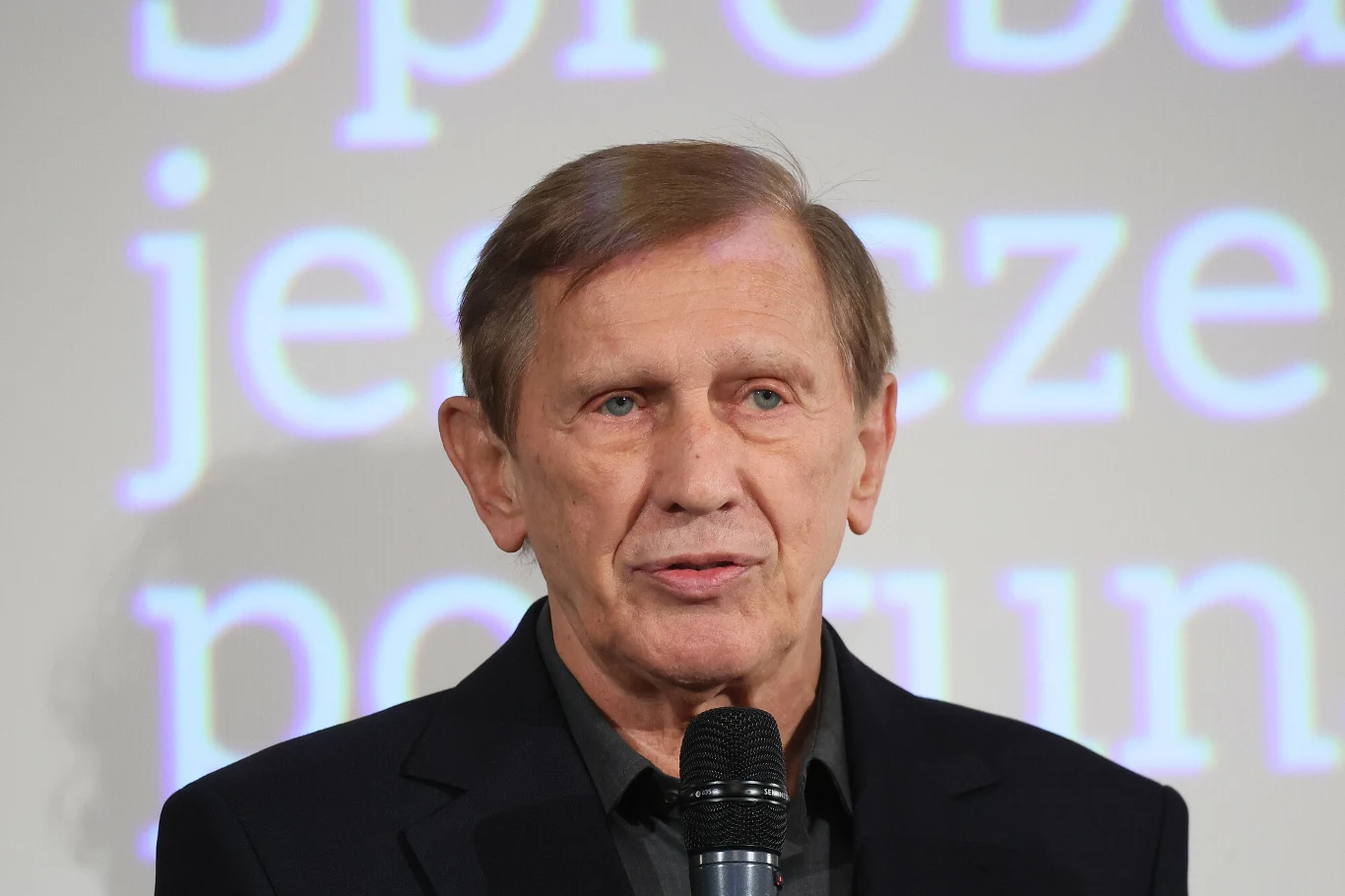 Jan Englert Mężczyzna w średnim wieku trzyma mikrofon, stoi na tle jasnego ekranu z dużymi, rozmytymi literami.