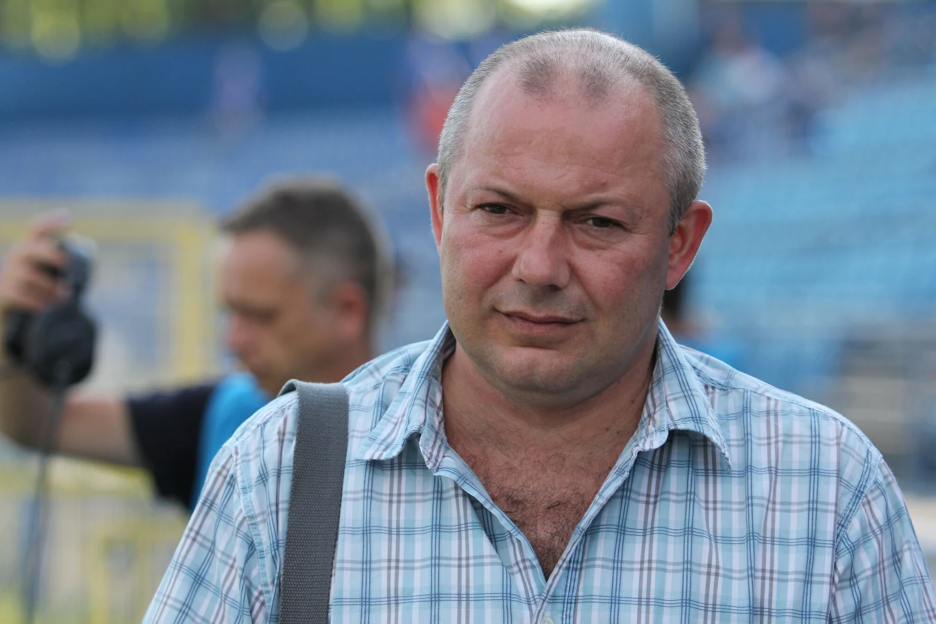 Mężczyzna w koszuli w kratę z krótkimi włosami, patrzący w stronę obiektywu, trzyma torbę na ramieniu, w tle rozmyty mężczyzna z aparatem oraz trybuny stadionu.