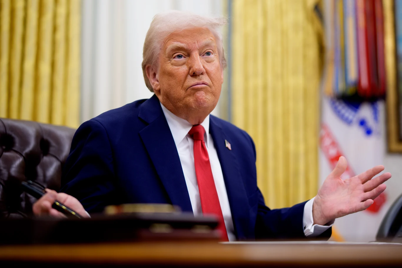 Donald Trump zmienił zdanie w sprawie nałożenia 25-procentowych ceł na samochody produkowane w UE Donald Trump w garniturze siedzi przy biurku w gabinecie z zasłonami i flagami w tle, gestykuluje dłońmi.