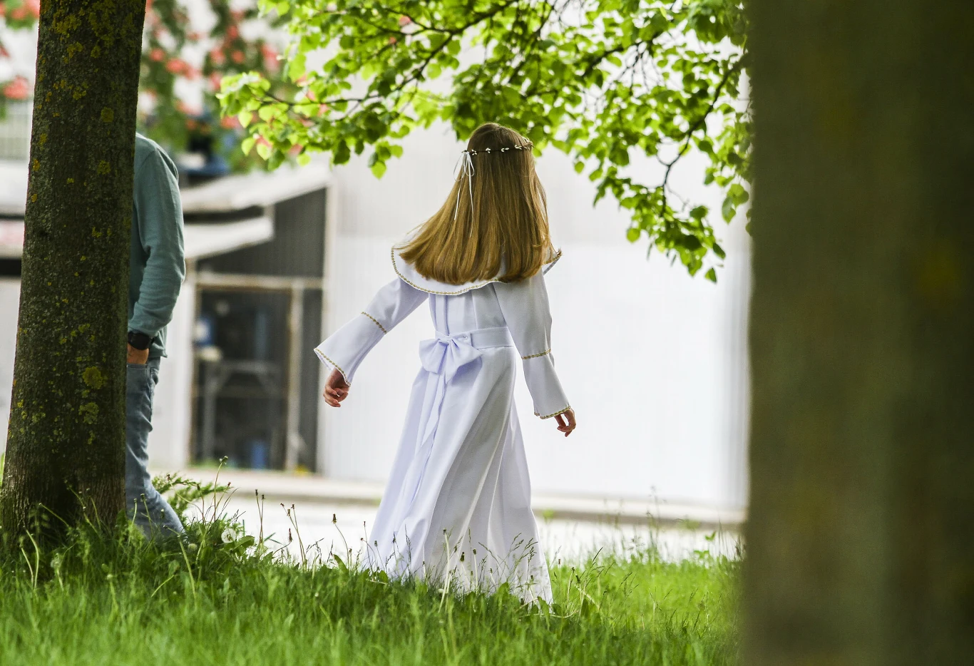 Dziewczynka w białej sukience komunijnej idąca pośród drzew, z długimi, rozpuszczonymi włosami ozdobionymi wiankiem.