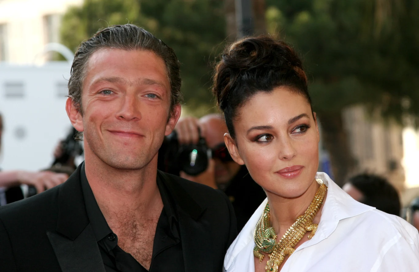 Vincent Cassel i Monica Bellucci Para elegancko ubranych osób pozuje razem, mężczyzna w ciemnym garniturze uśmiecha się lekko, kobieta w białej koszuli ozdobionej złotym naszyjnikiem patrzy w bok.