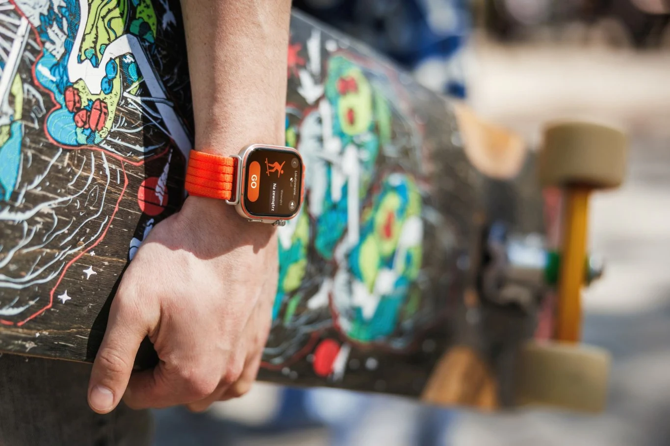 Dłoń trzymająca deskorolkę z kolorową grafiką, na nadgarstku pomarańczowy smartwatch z wyświetlaczem.