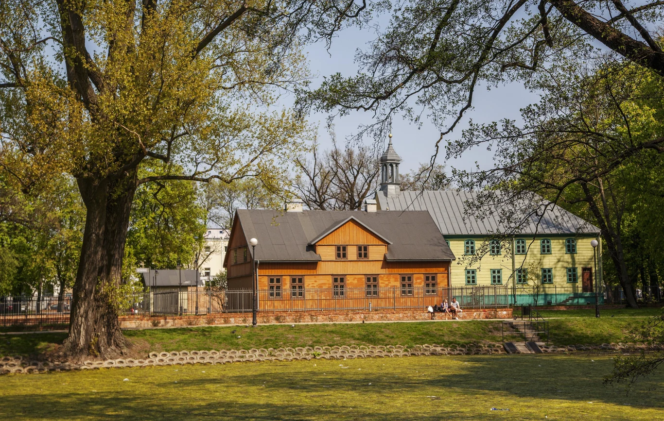 Drewniany dom o ciemnym dachu otoczony dużymi, rozłożystymi drzewami, obok pastelowego budynku z wieżyczką, na trawniku widoczne ławki i dwoje siedzących ludzi.