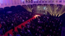 Duża sala wypełniona publicznością skierowaną w stronę oświetlonej sceny, na której trwa gala wręczenia nagród, a jasne reflektory i multimedialny ekran podkreślają centralny punkt wydarzenia.