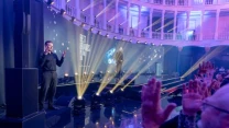 Mężczyzna przemawia ze sceny podczas gali, na której zgromadzona publiczność reaguje aplauzem, a całość rozświetlają efekty świetlne i elegancka oprawa wnętrza z balkonem.