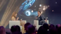 Kobieta w złotej sukni prowadzi rozmowę z mężczyzną w garniturze, oboje siedzą na białych fotelach na scenie, w tle duża grafika przedstawiająca muszlę z perłą oraz efekty świetlne.