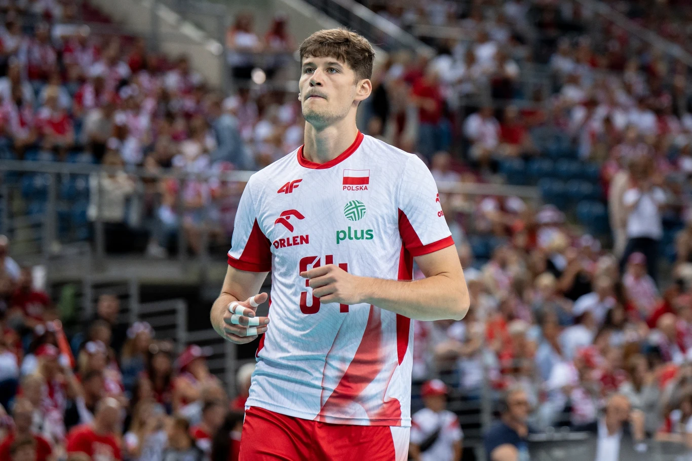 Siatkarz w koszulce reprezentacji Polski z numerem 6 stoi na hali sportowej, wokół widownia ubrana w biało-czerwone barwy narodowe.