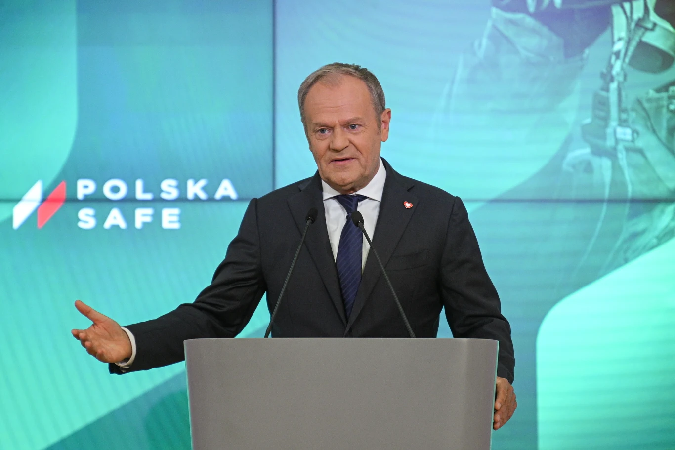 Mężczyzna w garniturze przemawia zza mównicy na tle ekranu z napisem „Polska Safe”.