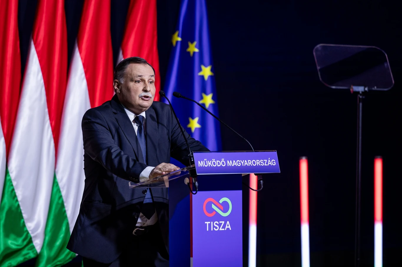 Mężczyzna w garniturze przemawia przy mównicy z napisem „TISZA”, w tle widoczne flagi Węgier oraz Unii Europejskiej.
