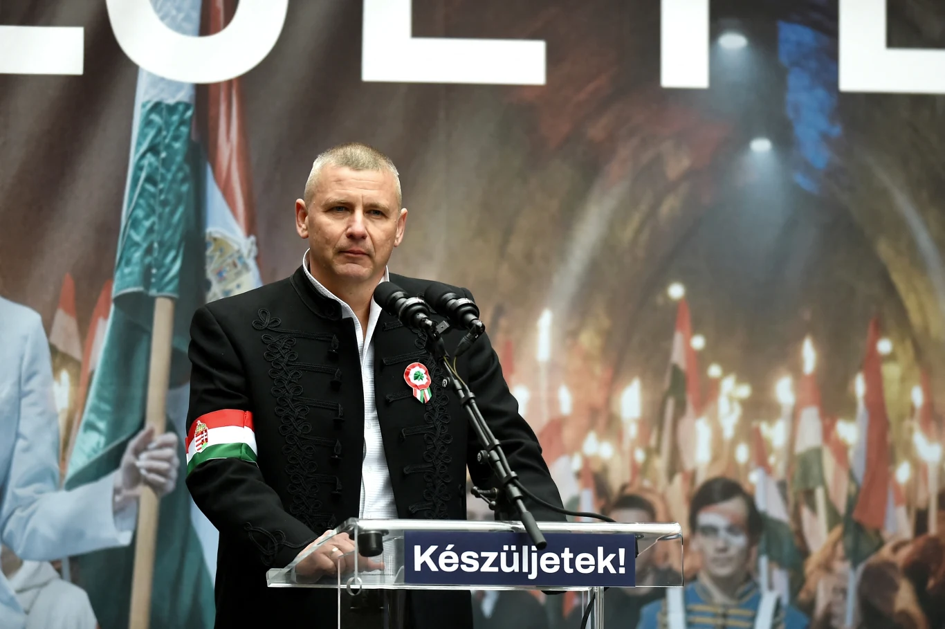 Mężczyzna w czarnym stroju z biało-czerwoną opaską na ramieniu stoi przy mównicy, za nim tłum ludzi trzymających w rękach węgierskie flagi oraz pochodnie, a na mównicy widoczny jest napis „Készüljetek!”.