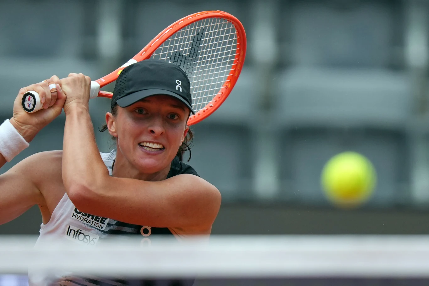 Tenisistka w czarnej czapce skupiona na odbiciu nadlatującej piłki, wyprowadzając mocny backhand dwuręczny na korcie.
