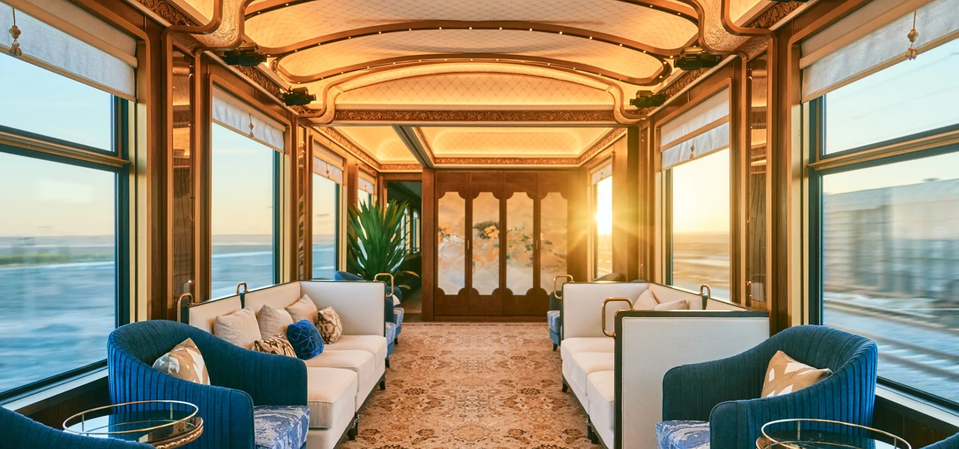 Luksusowy wagon pociągu z eleganckimi, niebiesko-białymi fotelami i sofami, bogato zdobionym sufitem oraz dużymi oknami, przez które wpada światło zachodzącego słońca.
