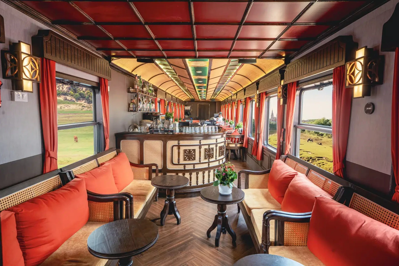 Luksusowy wagon restauracyjny pociągu z eleganckim barem pośrodku, wygodnymi sofami z pomarańczowymi poduszkami, niskimi stolikami oraz dużymi oknami zapewniającymi widok na zielony krajobraz za oknem.