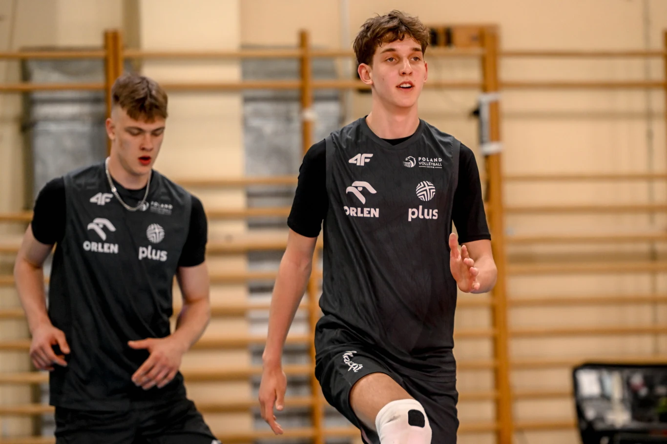 Dwóch młodych siatkarzy w sportowych koszulkach i spodenkach podczas treningu w sali gimnastycznej, jeden z nich wykonuje dynamiczny ruch, drugi podąża za nim.