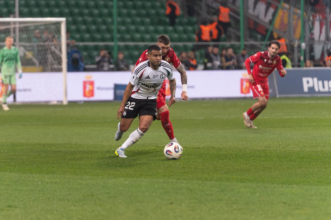 Mecz Legia - Widzew Piłkarz w białej koszulce prowadzi piłkę na boisku, naciskany przez zawodnika drużyny przeciwnej ubranej na czerwono, podczas meczu piłkarskiego na stadionie.