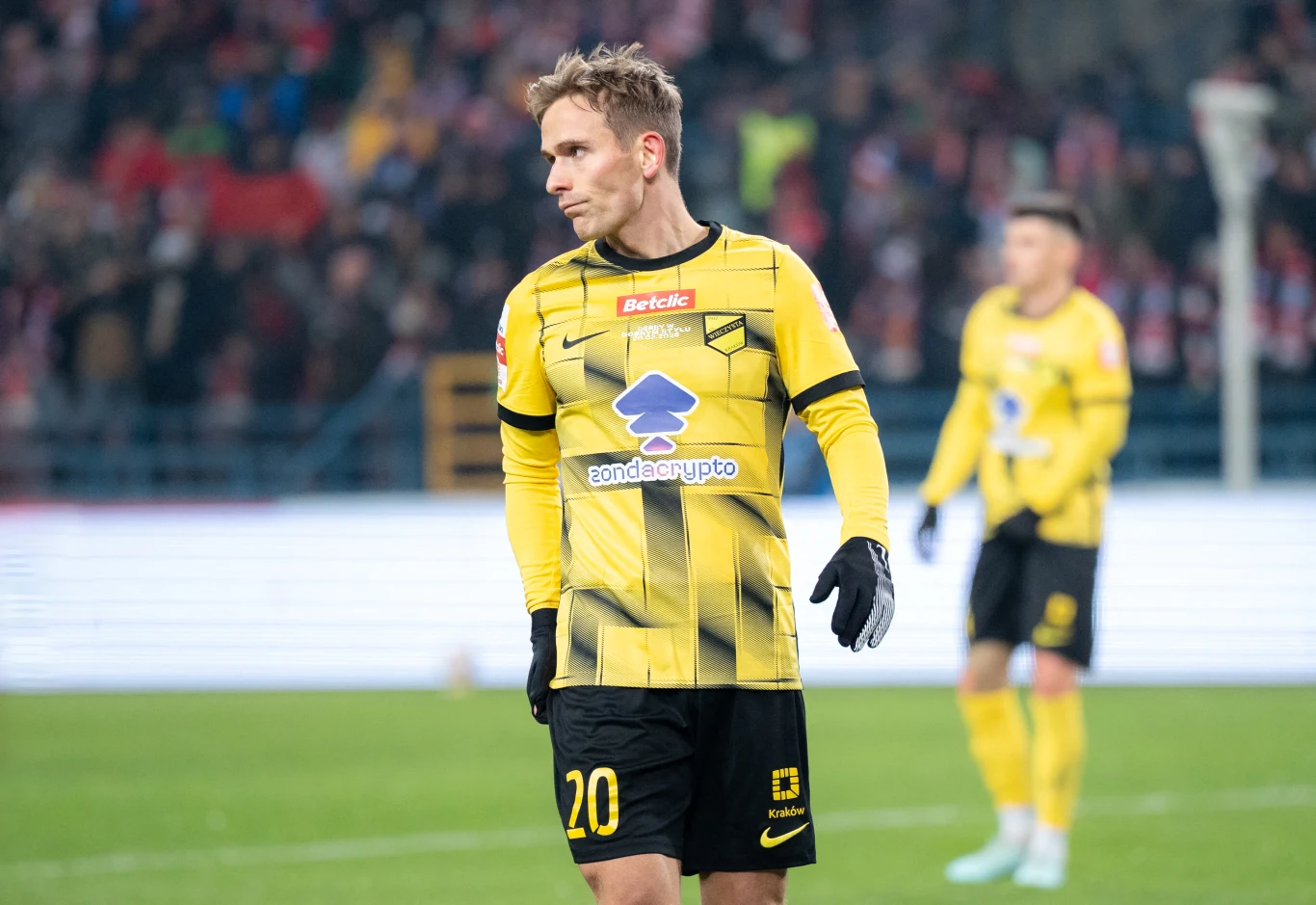 Mikkel Maigaard Piłkarz w żółto-czarnym stroju z numerem 20 stoi na murawie podczas meczu piłki nożnej, w tle widoczny inny zawodnik tej samej drużyny.