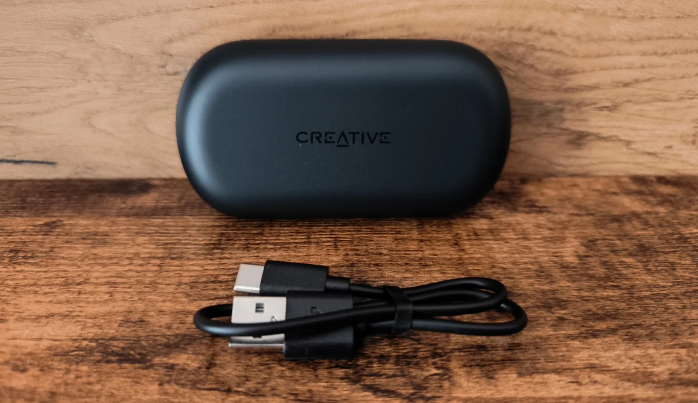 Czarne etui marki Creative ustawione na drewnianym blacie, przed nim ułożony czarny kabel USB z dwoma różnymi końcówkami.