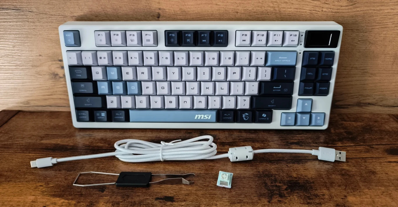 Mechaniczna klawiatura MSI z wielokolorowymi klawiszami, odłączanym białym kablem USB-C, narzędziem do zdejmowania klawiszy oraz podłączanym modułem i adapterem USB, ustawiona na drewnianym blacie.