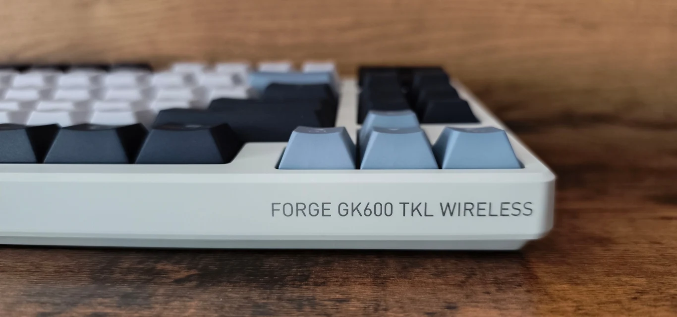 Klawiatura mechaniczna FORGE GK600 TKL WIRELESS z wyraźnie oznaczonym logo na obudowie, wyprofilowane, dwukolorowe klawisze na tle drewnianego blatu.