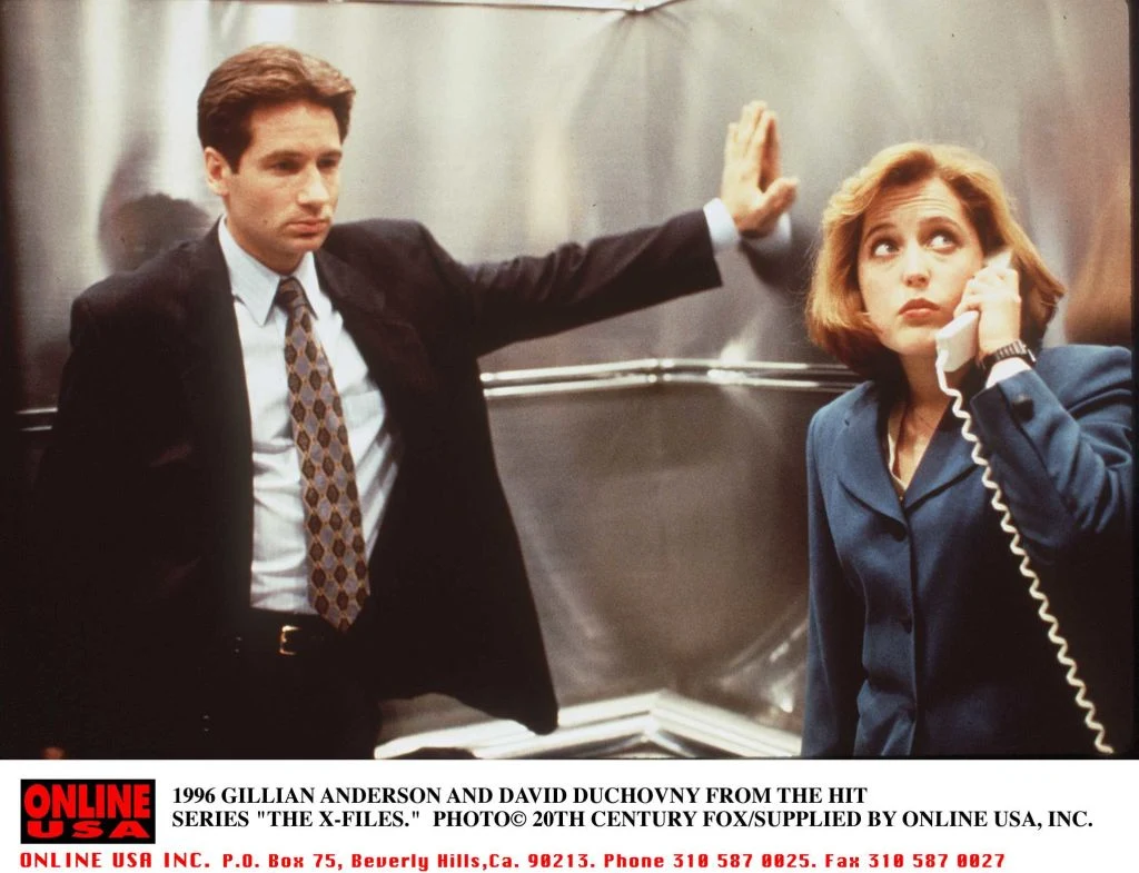 Gillian Anderson w serialu "Z Archiwum X" Mężczyzna w garniturze opiera się o ścianę windy, patrząc poważnie przed siebie, obok kobieta w kostiumie służbowym trzyma słuchawkę starego telefonu i spogląda w bok z zamyśleniem.