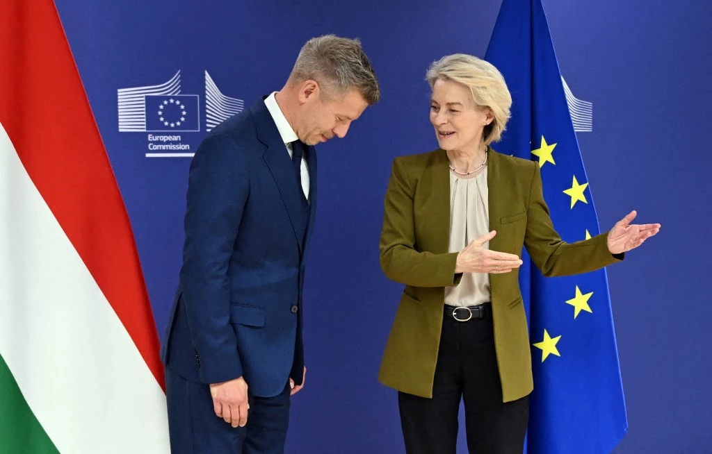 Dwoje elegancko ubranych polityków stoi obok siebie na tle flag Unii Europejskiej i Węgier; kobieta uśmiechnięta, gestykuluje w kierunku rozmówcy.