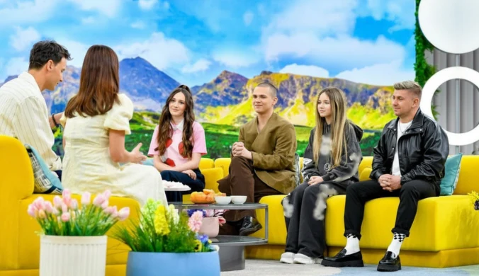Pięć osób siedzi na żółtych sofach w jasnym, nowocześnie urządzonym studiu telewizyjnym, za nimi krajobraz gór, na pierwszym planie wiosenne kwiaty w doniczkach.