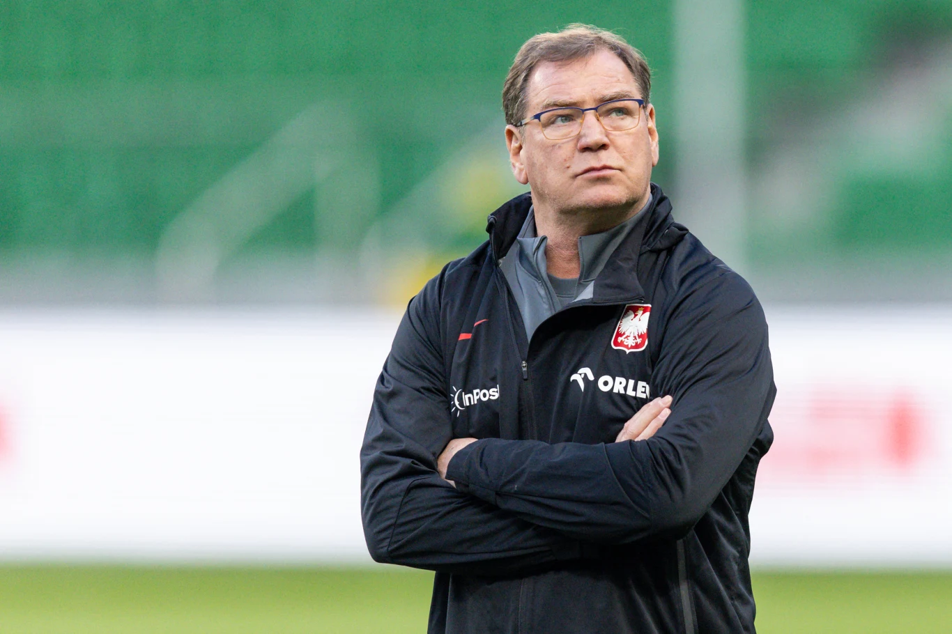 Mężczyzna w okularach, ubrany w czarną sportową kurtkę z logo polskiej reprezentacji piłkarskiej i napisem Orlen, stoi z założonymi rękami na tle zielonych trybun stadionu.