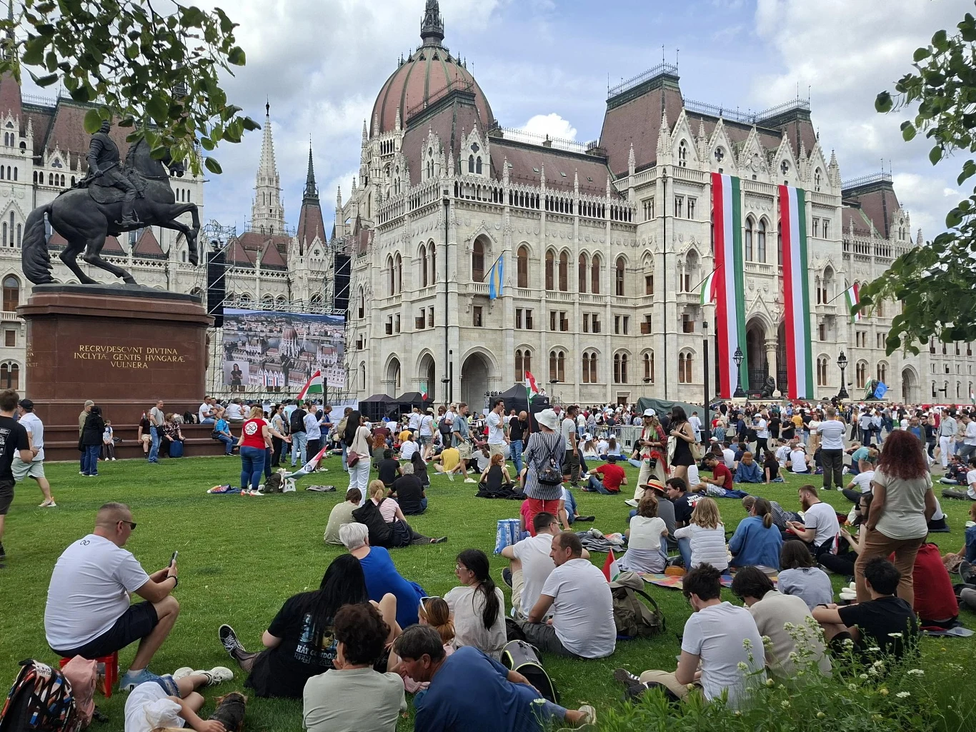Tłum ludzi siedzących i stojących na trawniku przed monumentalnym budynkiem parlamentu ozdobionym dużym pionowym pasem flagi Węgier, obok konny pomnik i scena koncertowa na tle miejskiej architektury.