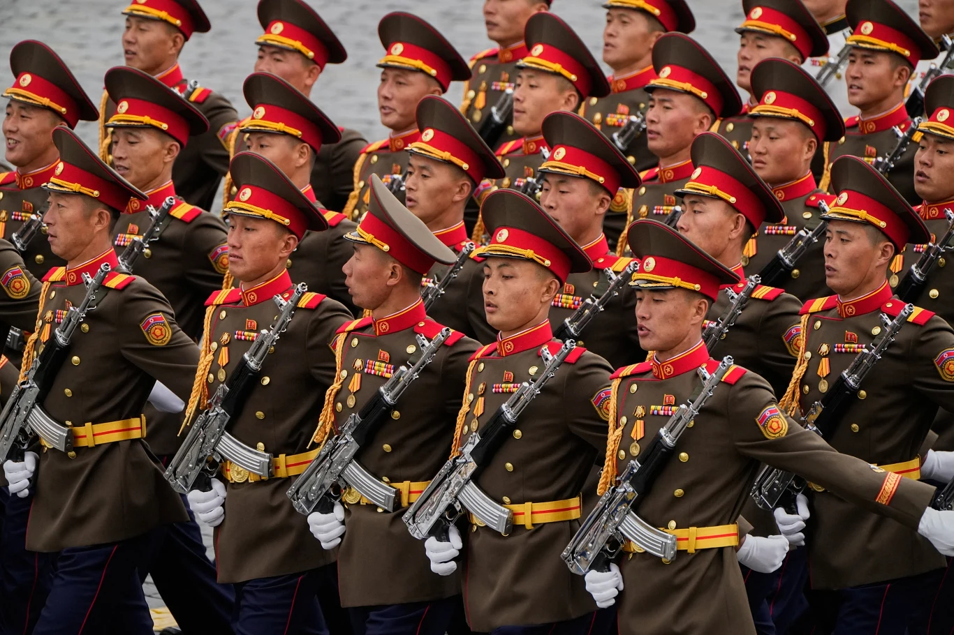 Rząd żołnierzy w mundurach wojskowych z czerwonymi czapkami maszeruje w szyku, każdy trzyma karabin na ramieniu i nosi liczne odznaczenia na piersi.