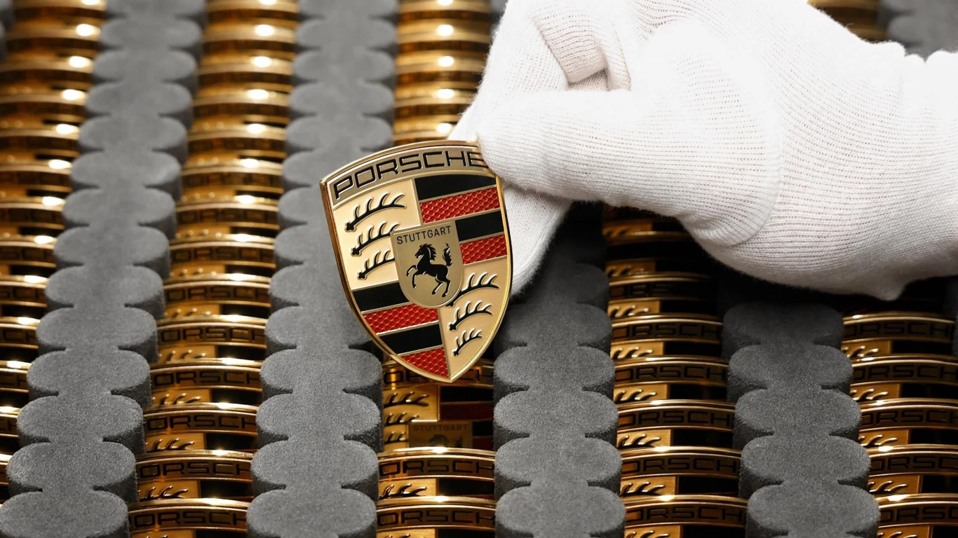 Porsche zmienia strategię, w efekcie pracę straci ponad 500 osób Biała rękawiczka trzyma emblemat Porsche na tle wielu złotych odznak marki ułożonych w rzędach na piance.